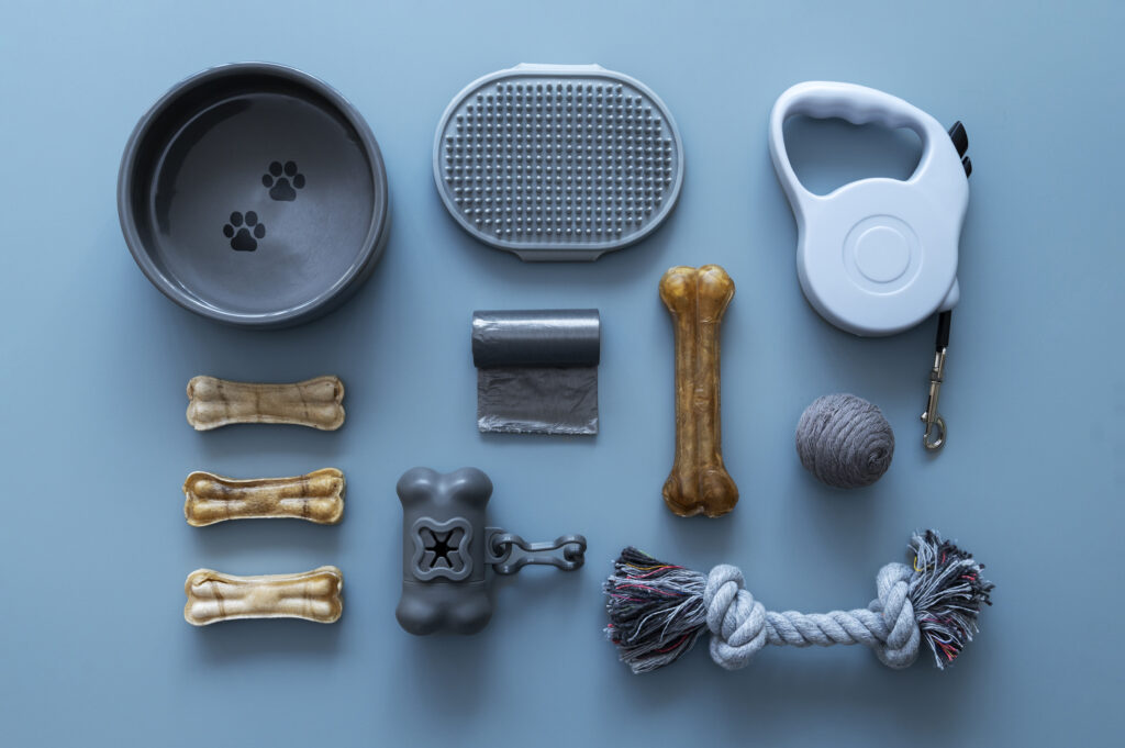 A flat lay of dog accessories, including a food bowl, bones, a leash, waste bags, and a rope toy, arranged neatly on a light blue background.