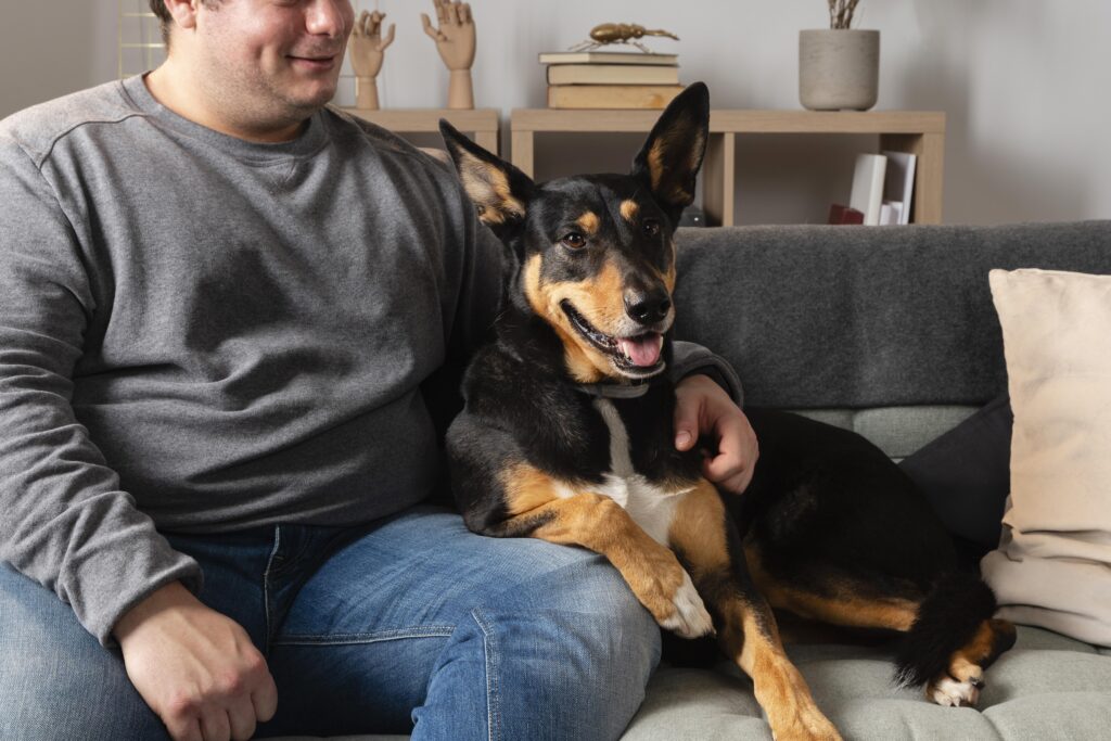 A man sitting on the couch with his dog, smiling at the camera