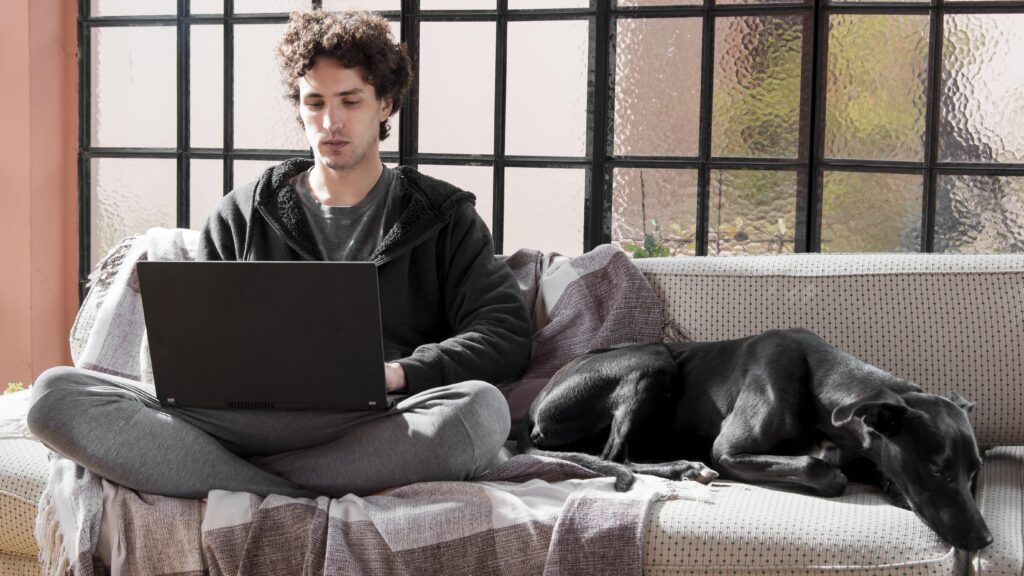 A man working on his laptop with a black dog resting beside him on the sofa