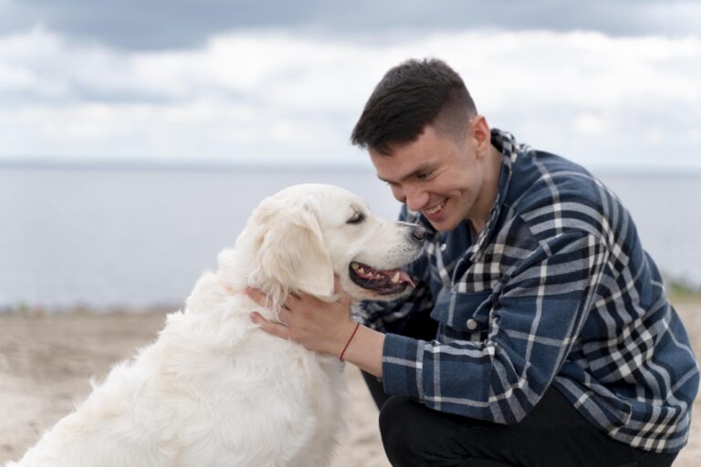 A man in a plaid shirt smiling warmly as he gently holds a fluffy white dog by the beach.