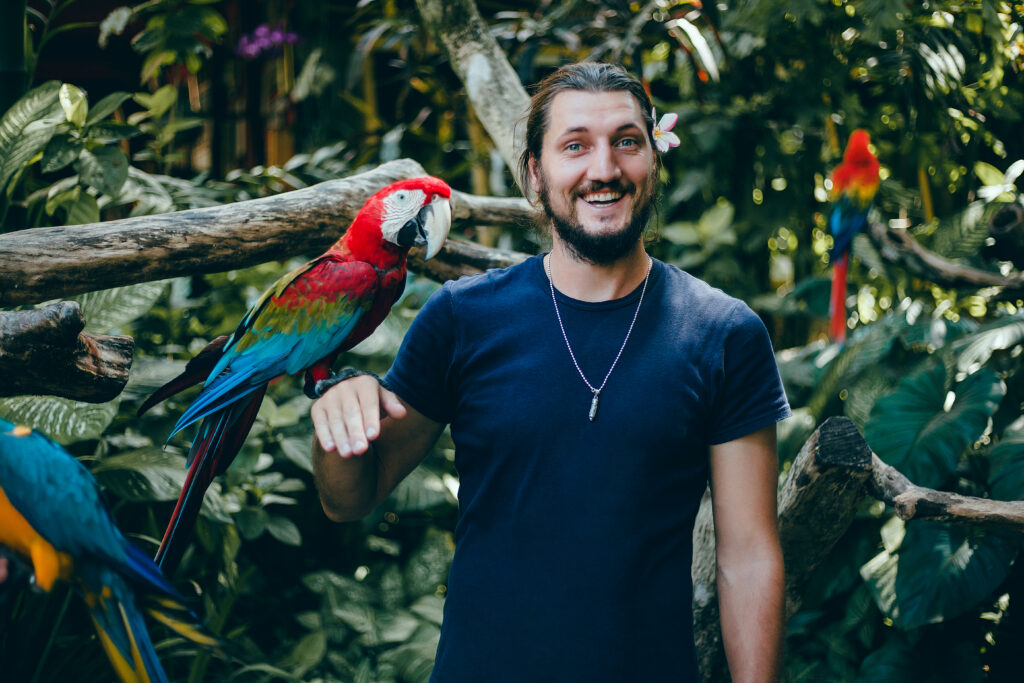 A man in a zoo smiling with a colorful parrot perched on his hand.